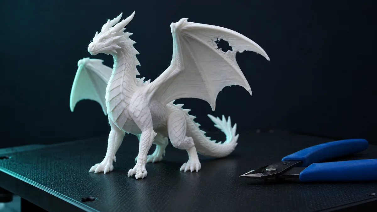 Dragão impresso em 3D com falha na asa sobre a mesa da impressora, alicate ao lado